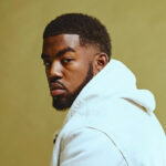 black man named tionwayne in front of a yellow wall with a white sweater looking into the camera